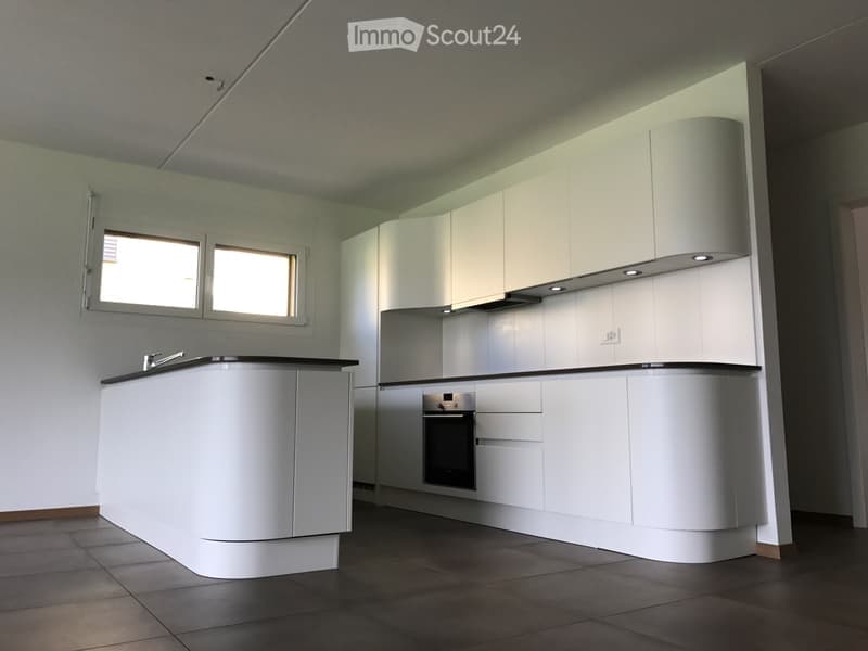 Modern kitchen with white cabinetry, dark countertops, and built-in appliances including a sink, oven, and range hood. The space features large windows and a clean, minimalist design.