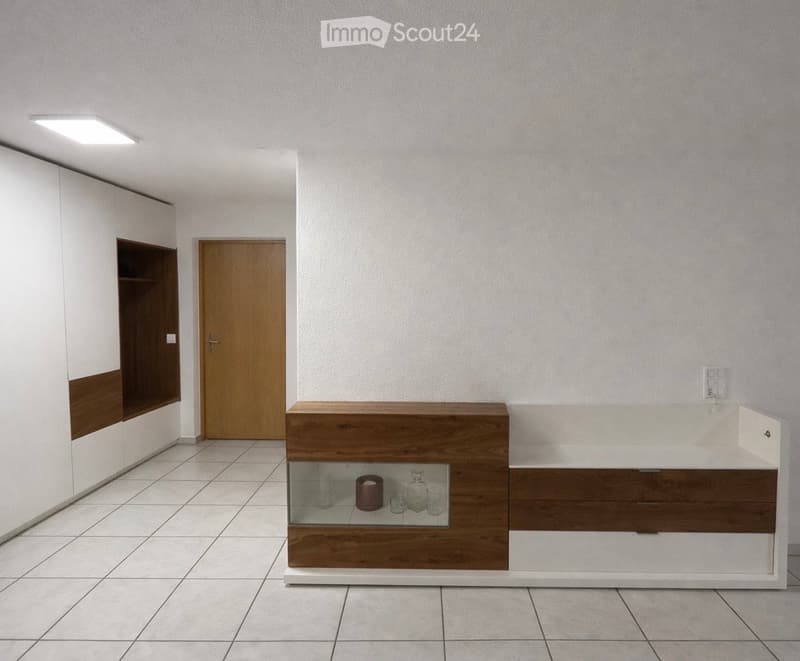 Empty room with tiled floor, white walls, a wooden door, and a built-in wooden bench with a glass display case containing decorative items.