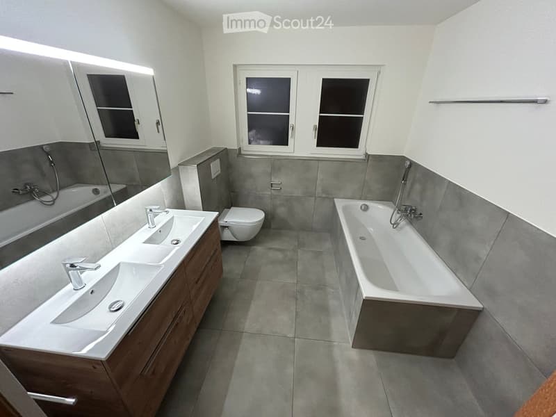 Modern bathroom with double sink, bathtub, toilet, and large mirror. Grey tiles and wooden cabinetry.
