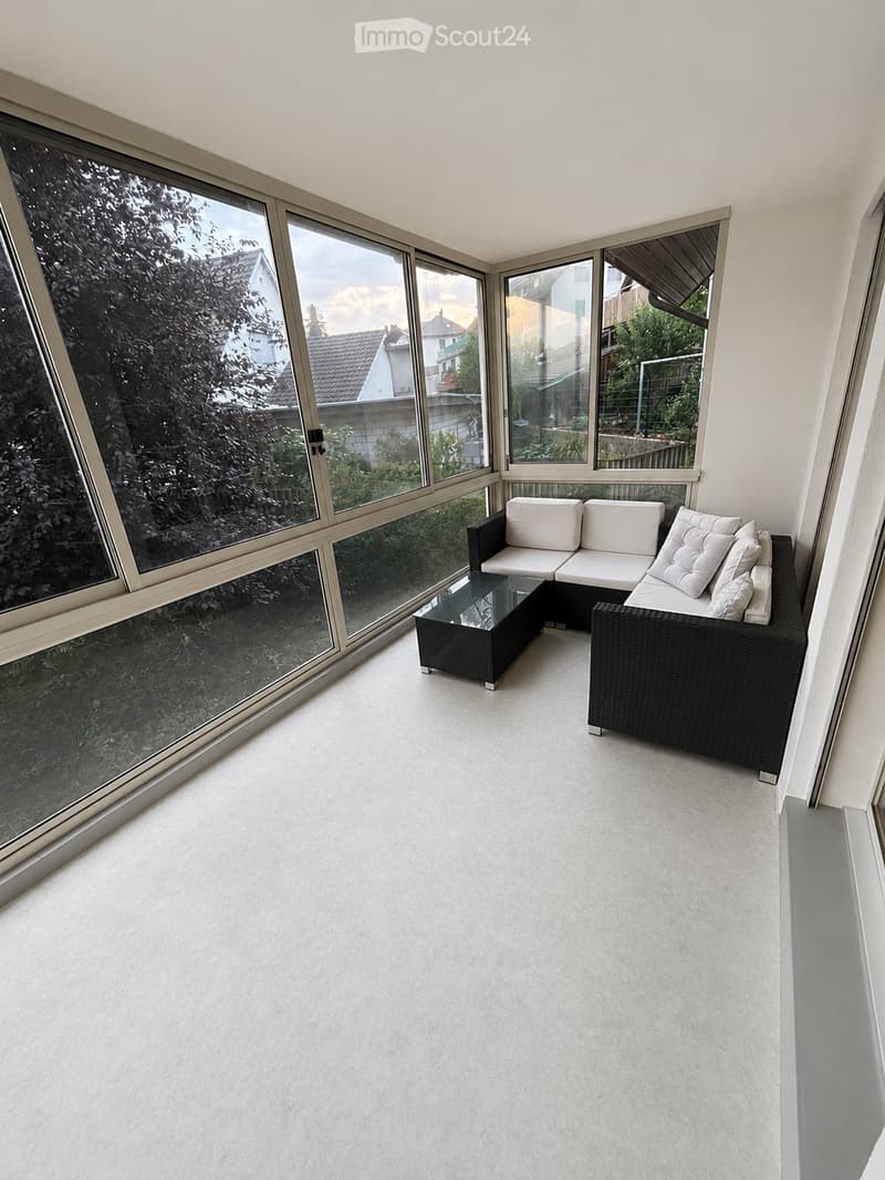 A modern, enclosed balcony with large glass windows, white carpeting, and a black wicker sofa set with a coffee table.