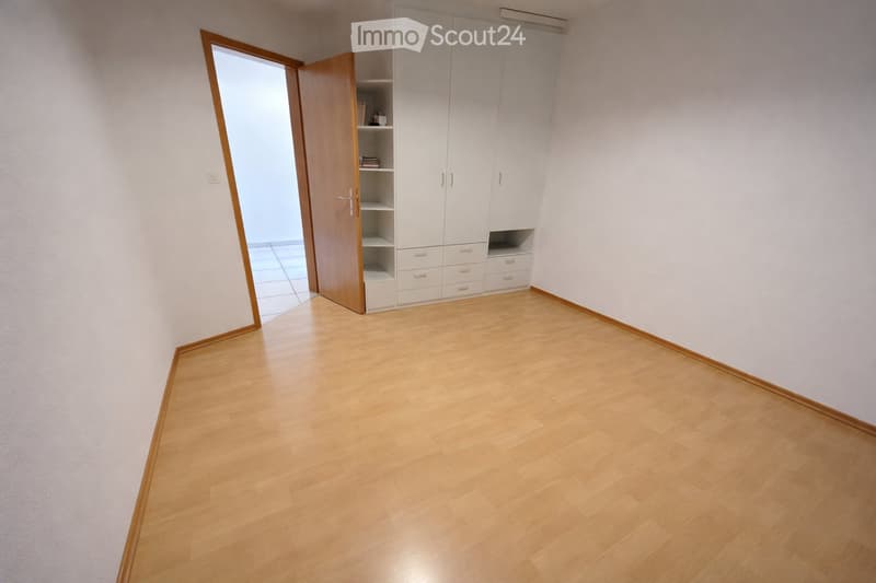 Empty room with light wooden flooring, white walls, and a built-in white wardrobe with shelves and drawers. The room has a wooden door leading to another space.