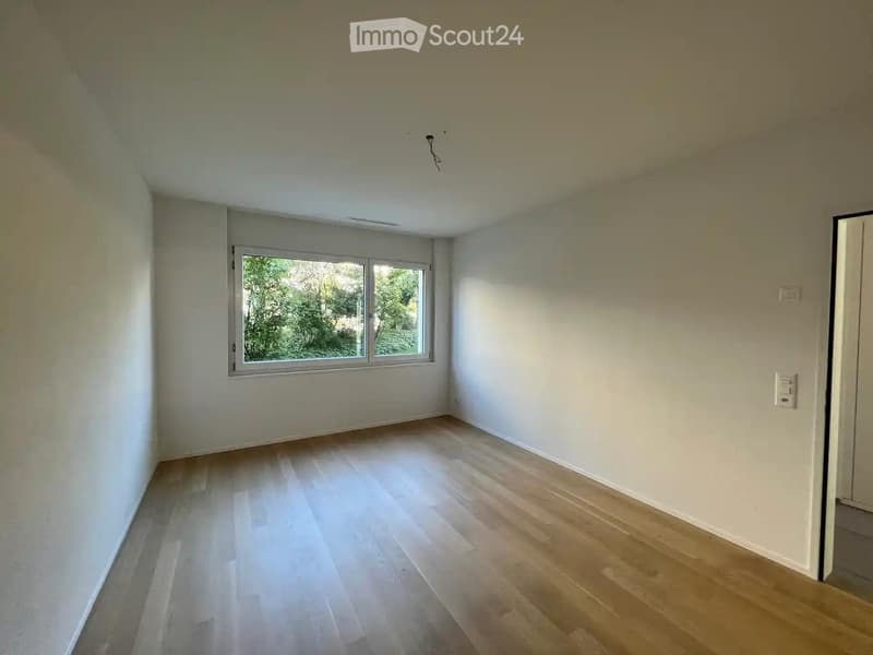 Empty room with light-colored wooden flooring, white walls, and a large window with a view of greenery outside.