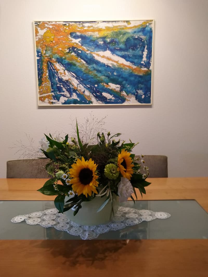 A glass table with a floral arrangement in a vase, placed on a white doily, with a wooden chair and a colorful abstract painting on the wall in the background.