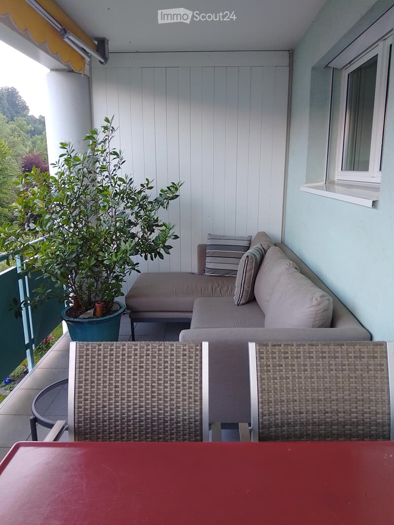 A small, modern balcony with a light blue floor, a white wooden slatted wall, a grey sofa with striped cushions, and a wicker basket. A potted plant adds greenery.