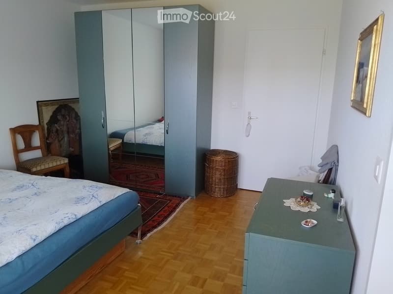 Bedroom with a wooden floor, a large bed with a blue and white duvet, a wooden chair, a large wardrobe with mirrored doors, a nightstand with decorative items, and a wicker basket.