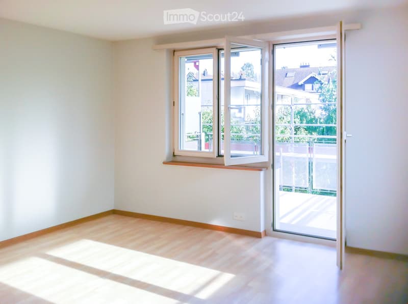 Empty room with light wooden flooring, white walls, and large windows opening to a balcony area.