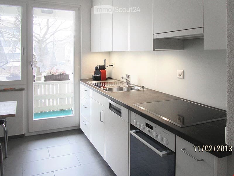 Modern kitchen with white cabinets, stainless steel sink, dishwasher, and coffee maker. Large window offers a view of the outdoor area.