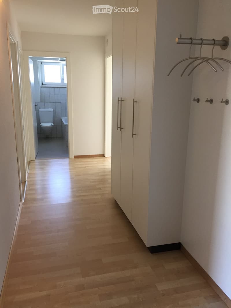 A clean, well-lit hallway with light wooden flooring, white walls, and a coat rack on the right side. A partially open door leads to a small bathroom with a toilet and sink visible.