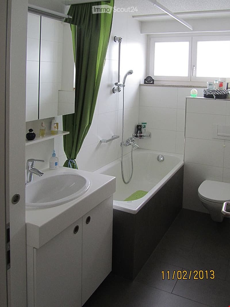 Modern bathroom with white tiles, a bathtub, a sink, a toilet, a mirror, and a green curtain. Shelves hold toiletries and a small clock is placed on the windowsill.