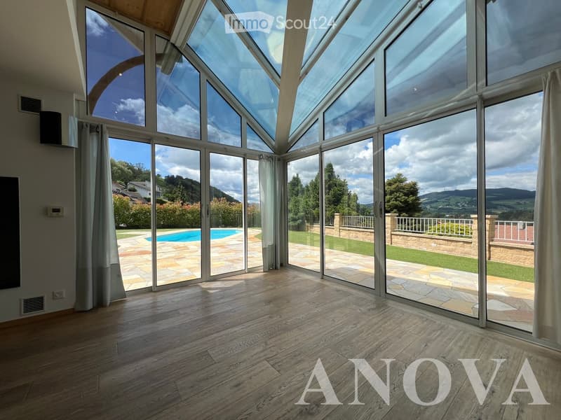 Bright, spacious room with large glass walls and doors opening to a patio and pool area. Wooden flooring, modern design, scenic mountain view.