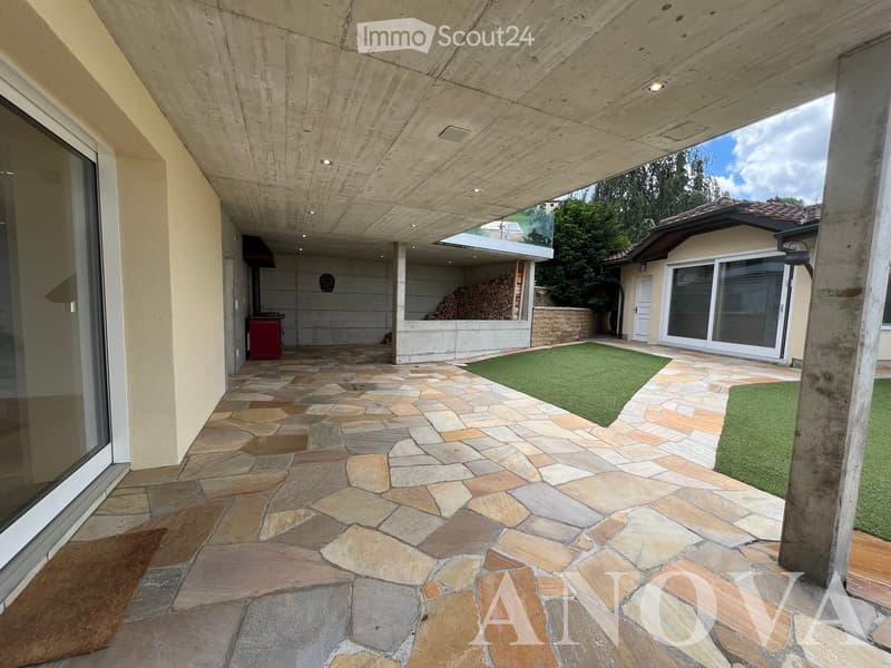 Outdoor patio with stone tiles, covered wooden ceiling, artificial grass patches, and a view of a house with large windows and a stone wall.
