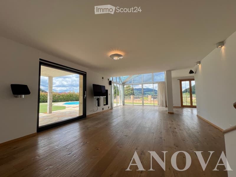 Bright, empty room with wooden flooring, large windows, and sliding doors leading to a patio and pool area with a scenic mountain view.