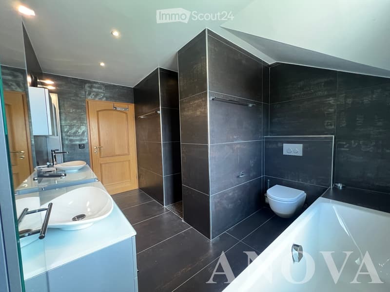 Modern bathroom with dark grey tiles, two sinks, a bathtub, and a toilet. Features include a large mirror, sleek faucets, and recessed lighting.