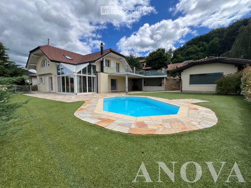 Lush green lawn with a swimming pool surrounded by stone tiles, modern house with large windows and a balcony, and a scenic mountain backdrop.