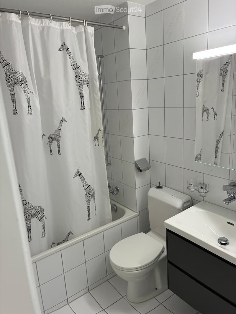 Modern bathroom with white tiles, a bathtub, toilet, sink, and a shower curtain featuring giraffe designs.