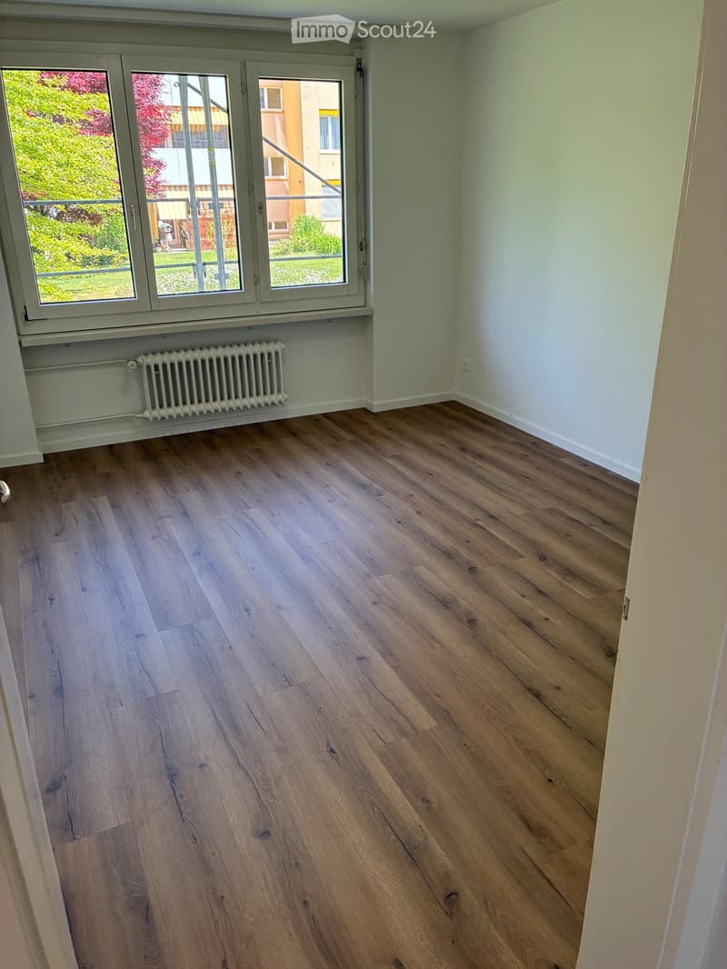 Empty room with light wooden flooring, white walls, and a large window with a radiator below it.
