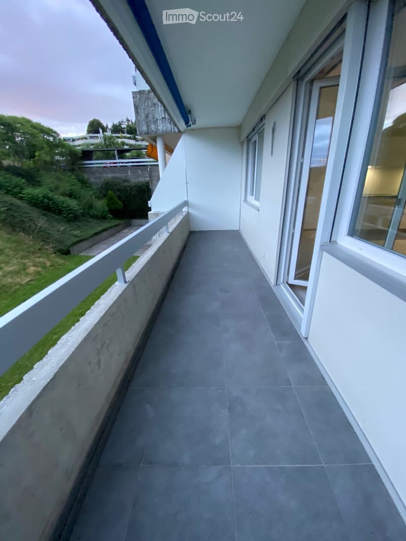 Empty balcony with grey tiled floor, white railing, and large windows overlooking a green area.