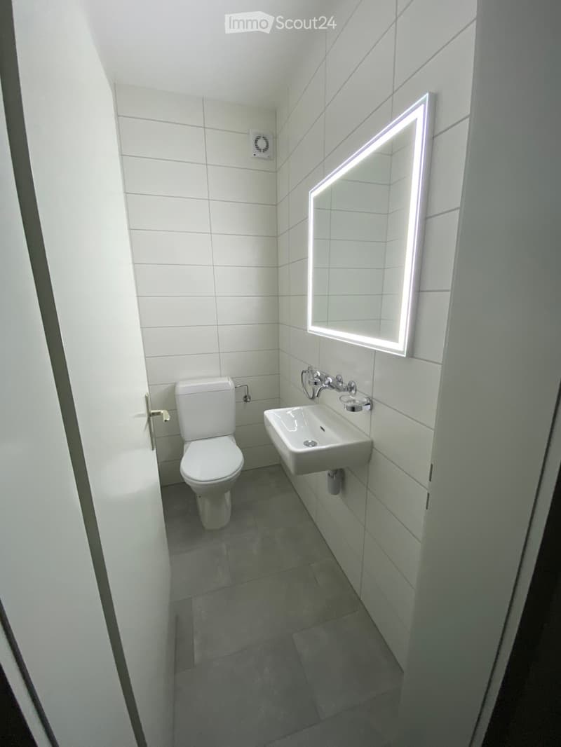 Modern bathroom with white tiled walls, a toilet, a sink, and a illuminated mirror above the sink.