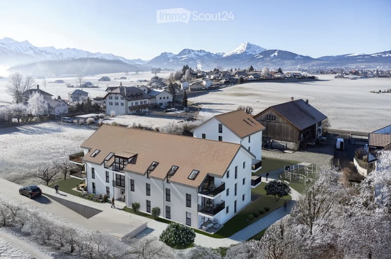 Aerial view of a modern two-story house with a brown tiled roof, white exterior, and multiple balconies. The house is surrounded by a landscaped garden with trees, a patio, and outdoor seating. The surrounding area includes other houses, a snow-covered fi