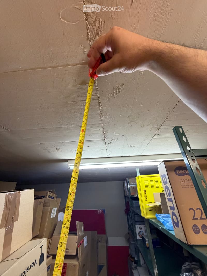 An unfurnished room with exposed ceiling beams, cardboard boxes, and a shelf with storage items. A hand is measuring a crack in the wall with a tape measure.