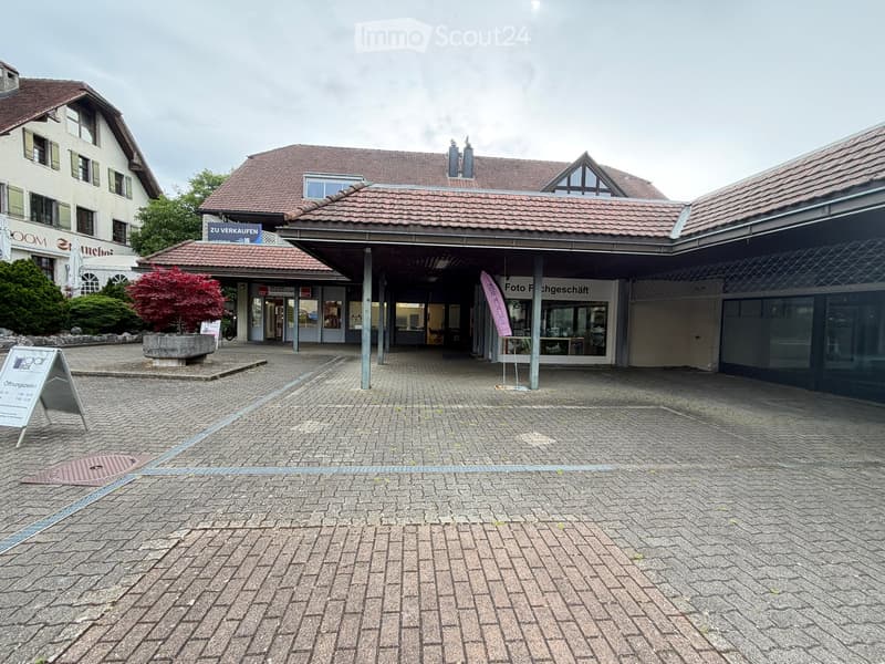 A paved courtyard area with a modern building entrance, a small tree, and signage for a photo shop.