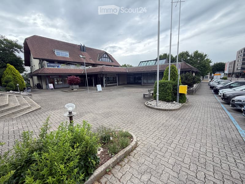A commercial building with a tiled roof and signage reading "ZU VERKAUFEN" (For Sale). The entrance features glass doors and a paved area with several parked cars and greenery.