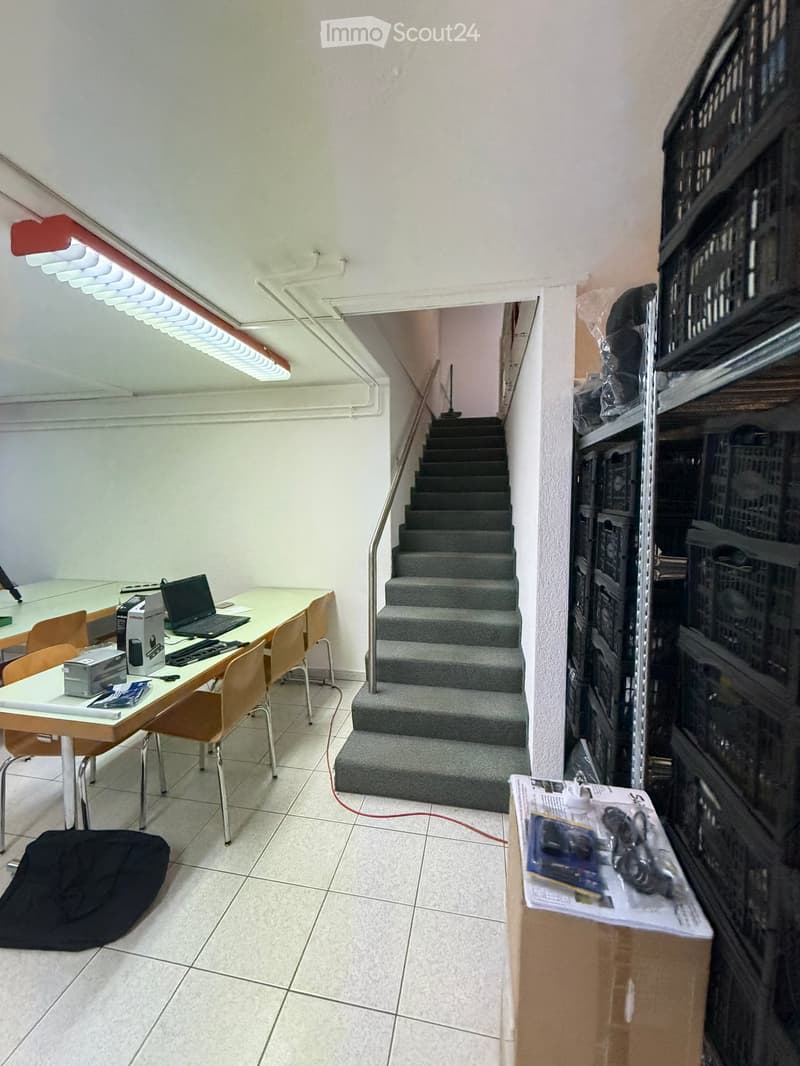 A furnished room with a desk, laptop, chairs, printer, and a beanie bag, leading to a staircase with a metal railing.