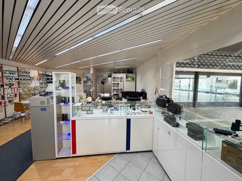 Modern retail space with a glass display counter, shelves filled with cameras and accessories, a small refrigerator, and a seating area with a chair and table.