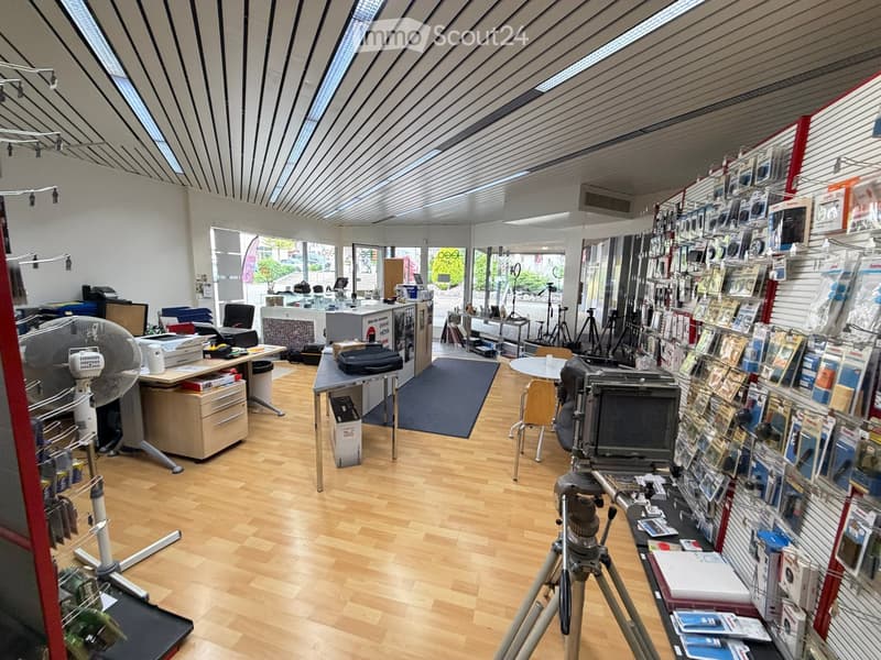 A well-lit electronics store with wooden flooring, large windows, and a variety of cameras, tripods, and shelves displaying accessories. There are desks, chairs, and a fan in the space.