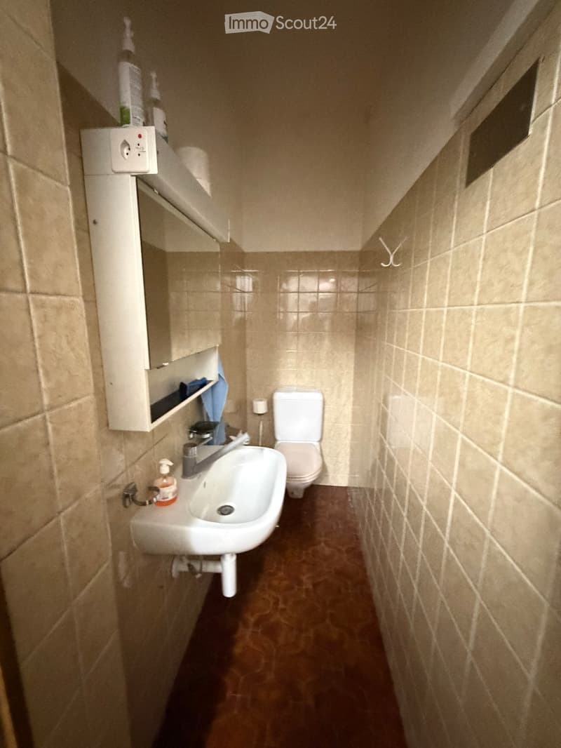 Small bathroom with beige tiled walls and brown tiled floor, featuring a white sink with a chrome faucet, a toilet, a mirrored cabinet, and a soap dispenser.