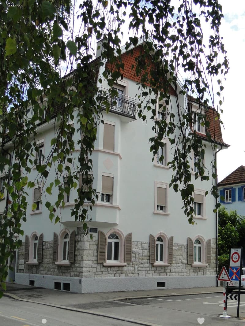 Three-story building with a red roof, white walls, brown trim, multiple windows, and a balcony on the second floor.