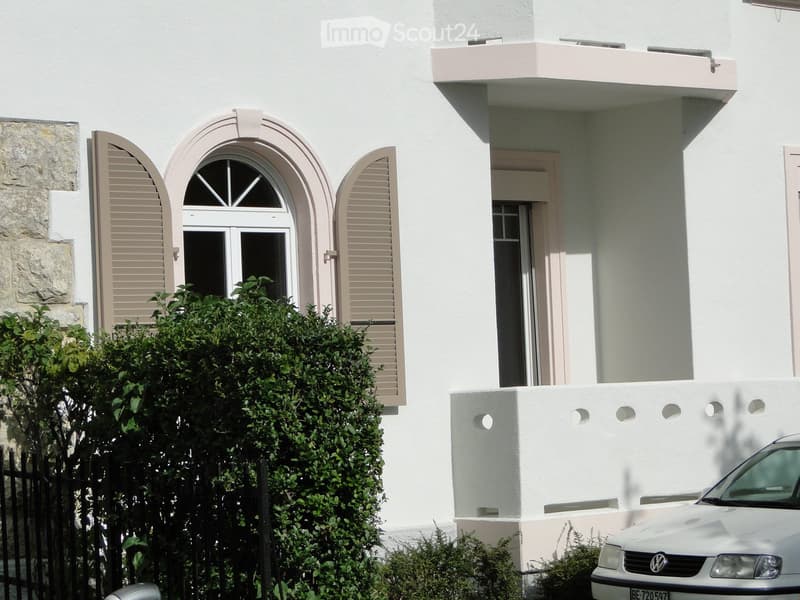 white painted house, stone wall on the left side, arched window, balcony, car parked in front