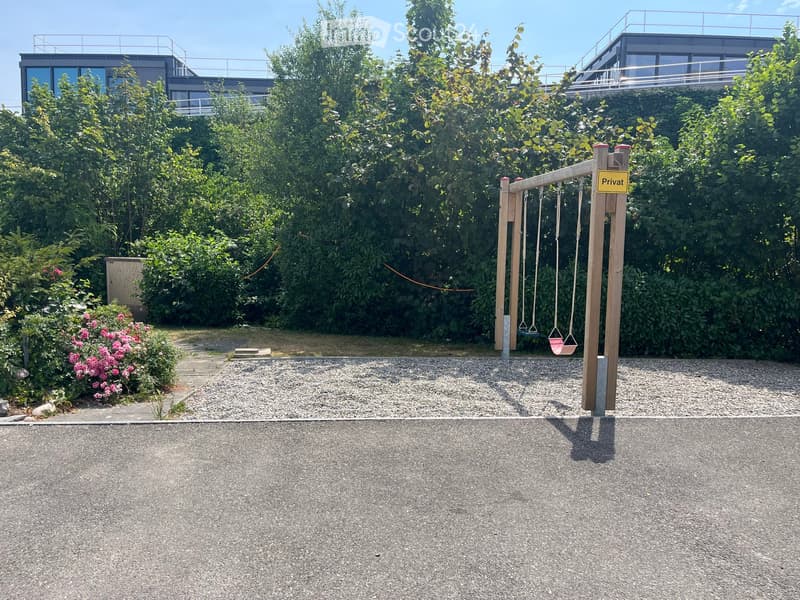 Private outdoor area with a swing set, surrounded by lush greenery and a modern building in the background.