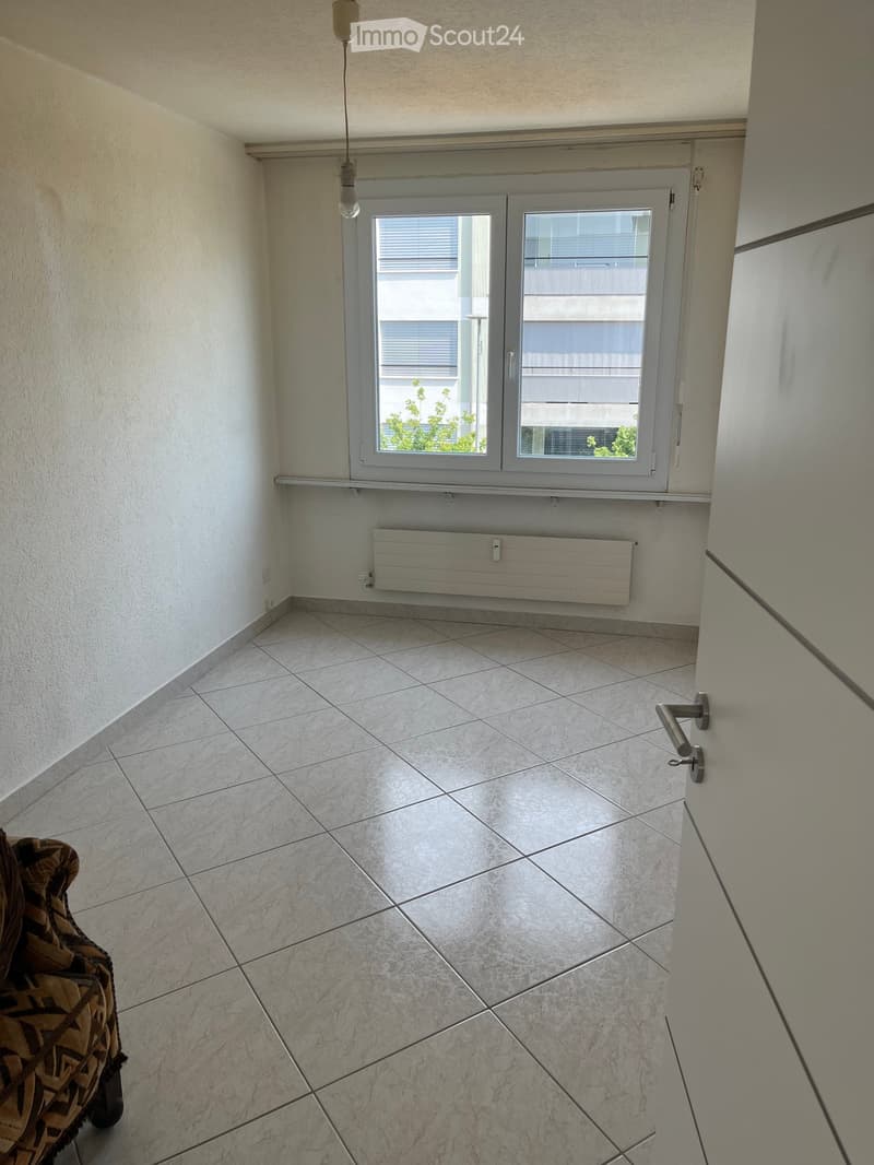 A small, unfurnished room with tiled flooring, a window with blinds, a radiator, and a single sofa partially visible on the left side.