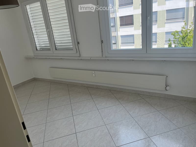 Empty room with tiled floor, white walls, large windows with blinds, and a radiator below the windows.