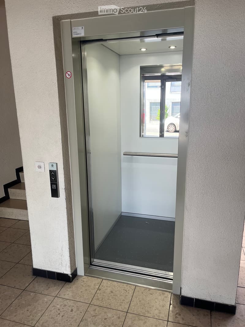 Elevator entrance in a hallway with tiled floor, control panel, and a view of the outside through the elevator door.