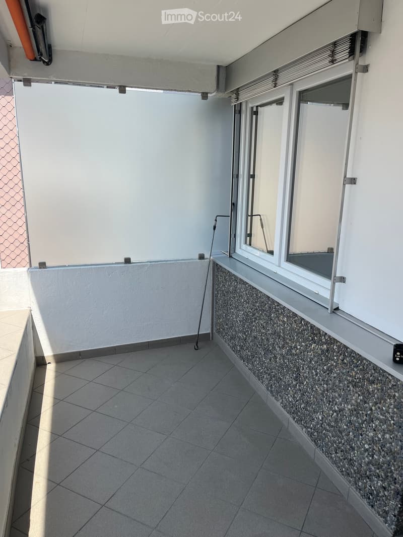 Balcony with tiled floor, white walls, and a small window with a view. The balcony has a low wall with a pebble finish along one side.
