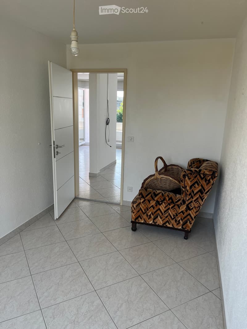 Hallway with tiled floor, white walls, a patterned sofa, and a wicker basket placed against the wall. An open door leads to another room.