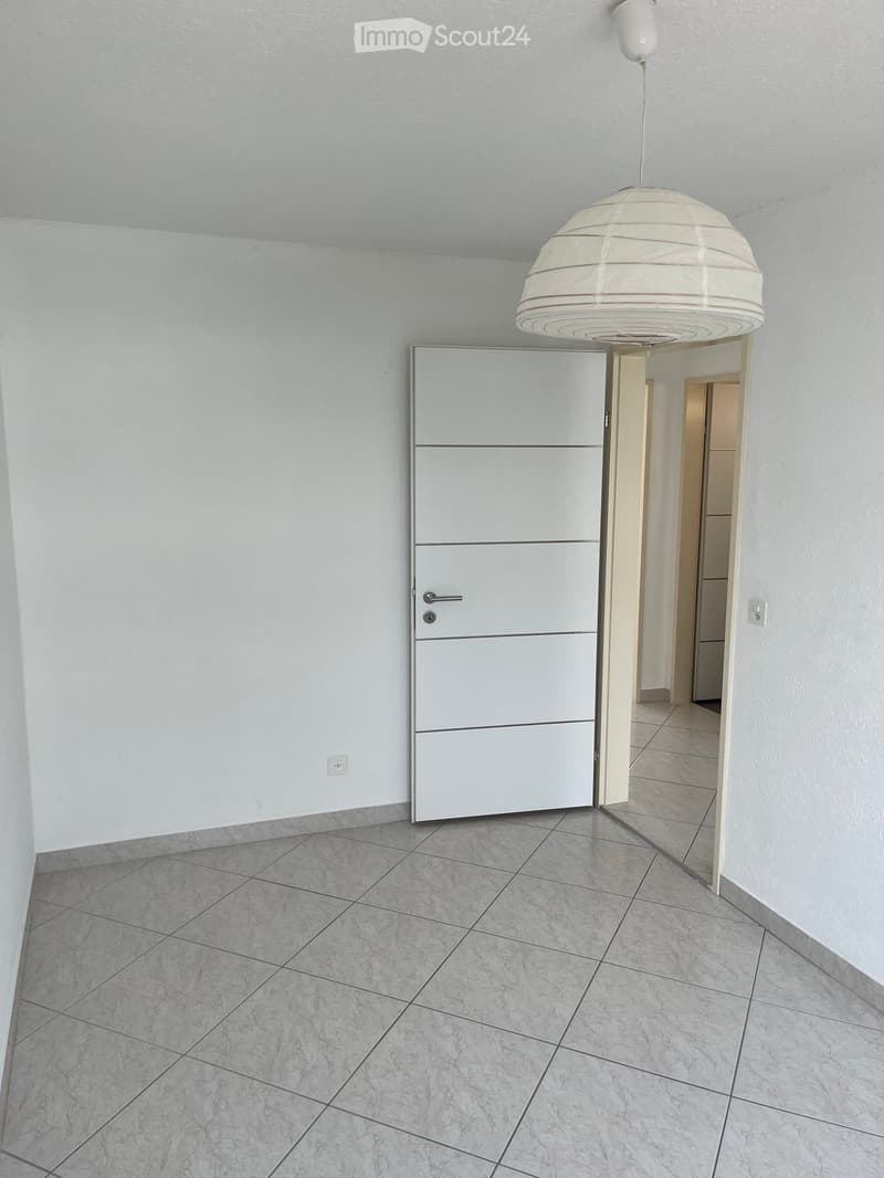 Empty hallway with white walls, tiled floor, and a modern white door leading to another room. A hanging light fixture is visible on the ceiling.
