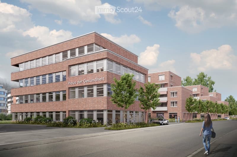 A modern brick building labeled "Haus der Gesundheit" with multiple floors, large windows, and balconies. The building is surrounded by greenery and a paved road.