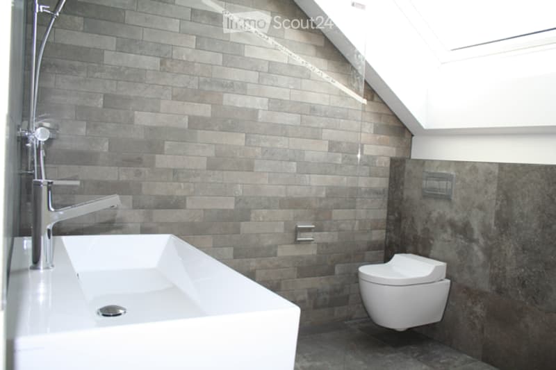 Modern bathroom with grey tiled walls, white sink, chrome faucet, and white toilet. The room features a skylight window and sleek, contemporary design.