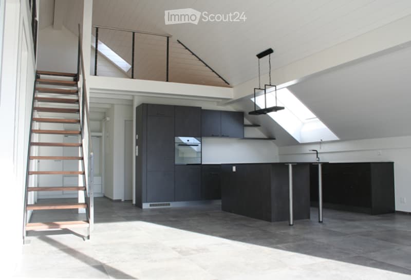 Modern kitchen with dark cabinets, a central island, and a staircase leading to an upper level. The space features a sleek design with a stove and stools at the island.