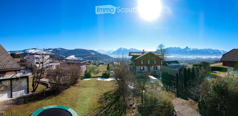 A panoramic view of a residential area with houses, green lawns, trees, and distant snow-capped mountains under a bright blue sky.