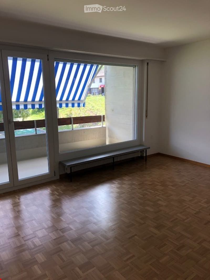 Large room with wooden floor, sliding glass doors, large windows with blue striped awnings, radiators, a bench, and a balcony with a view of a grassy area and a swimming pool