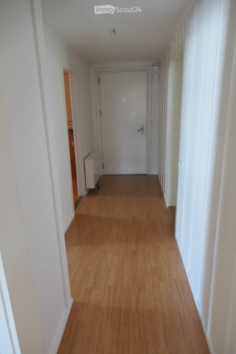 A narrow hallway with light wooden flooring and white walls, featuring a closed door at the end and a radiator on the left side.