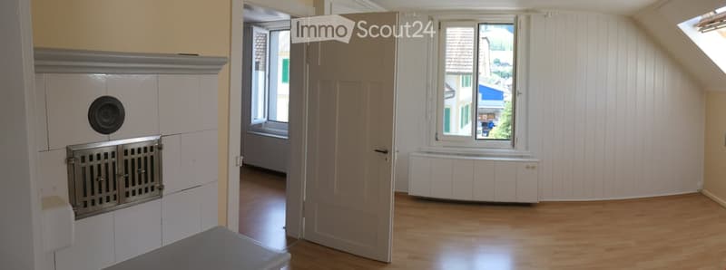 Empty attic room with wooden flooring, white paneled walls, and large windows offering a view of the outside. Features include a fireplace with a metal grate and a built-in bench beneath one of the windows.
