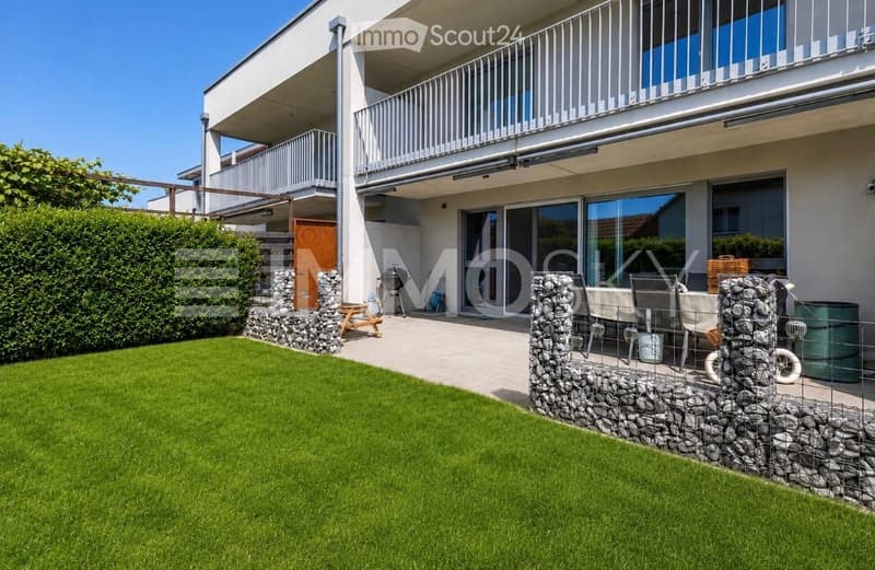 Well-maintained garden with a neatly trimmed lawn, stone walls, outdoor seating area, and a modern multi-story house with balconies in the background.