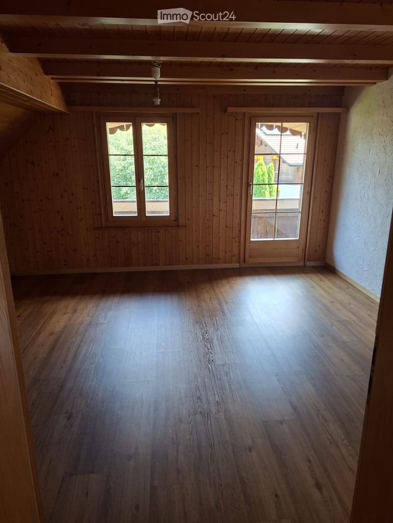Empty room with wooden walls and floor, two large windows, and a hanging light fixture.