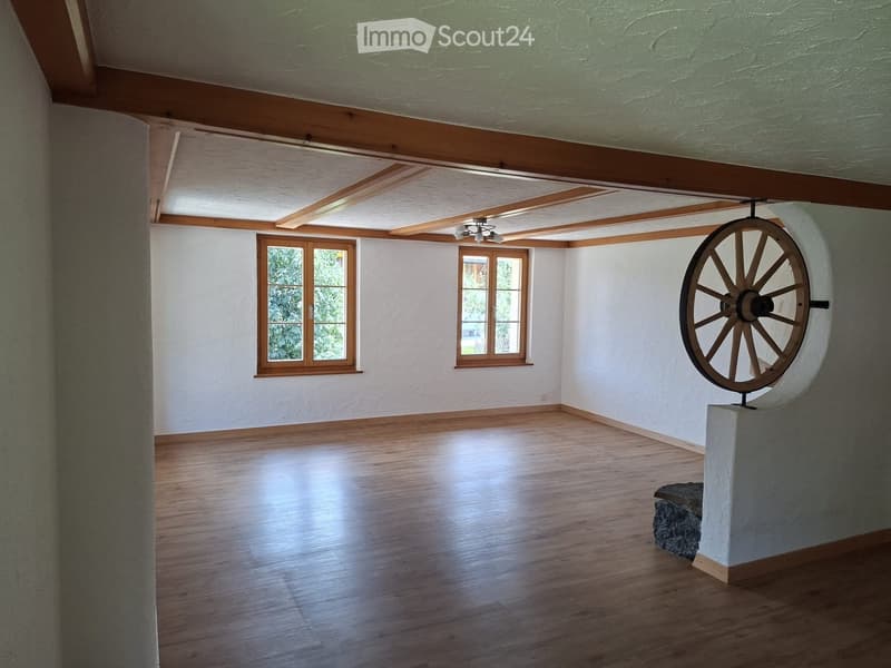 Empty room with wooden flooring, white walls, exposed wooden beams on the ceiling, and two windows with wooden frames. A large wooden wheel sculpture is placed on a pedestal in the corner.