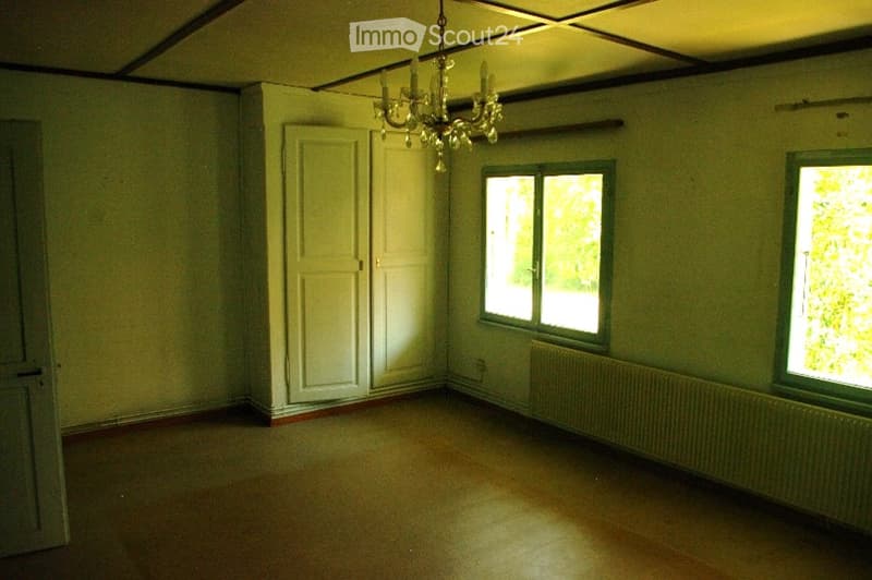 Empty room with wooden flooring, white walls, two windows, a door, and a chandelier hanging from the ceiling.